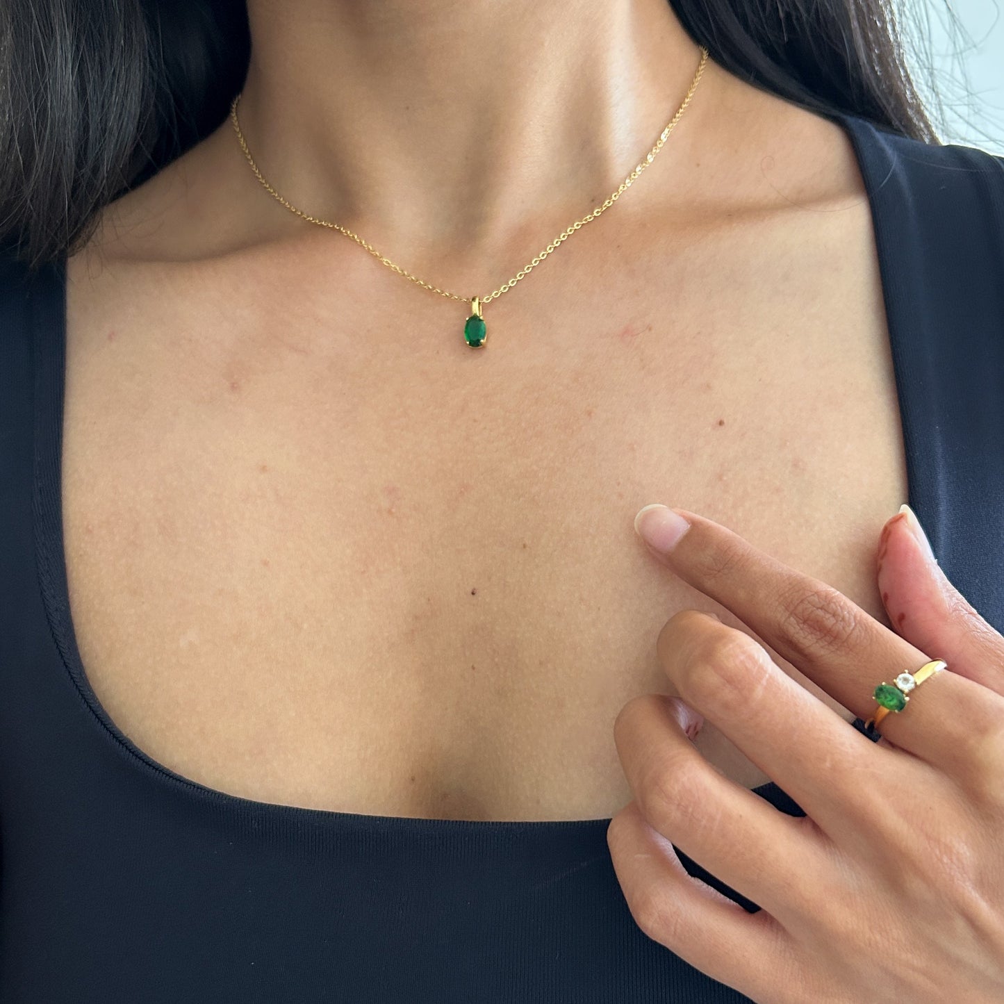 Woman wearing a gold necklace and ring with green gemstones, touching her chest.