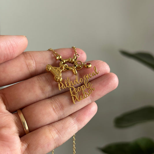 A gold necklace with a 'Pathological people pleaser' script pendant and a small animal charm, held in a person's hand against a soft-focus background.