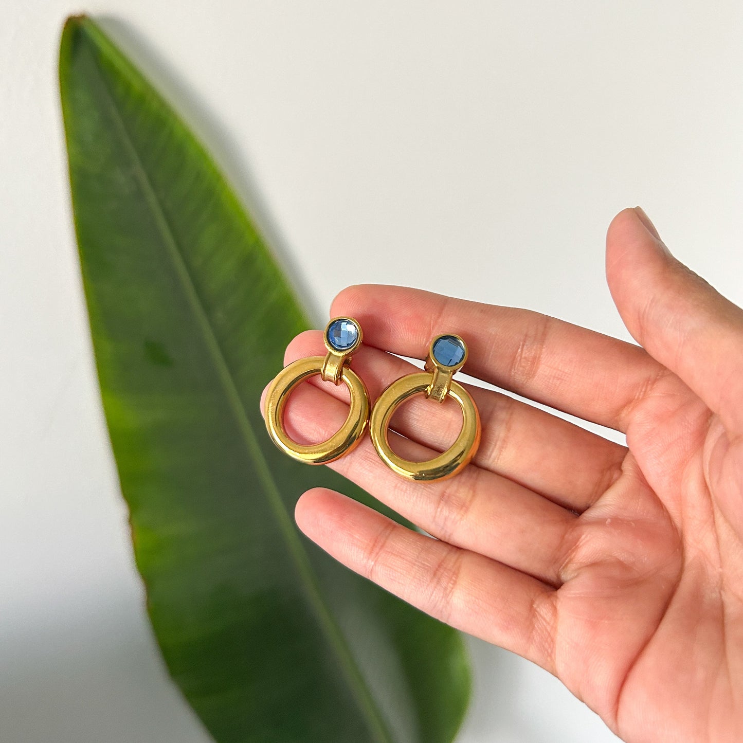 Gold earrings with blue gemstones held in a hand next to a green leaf on a white background