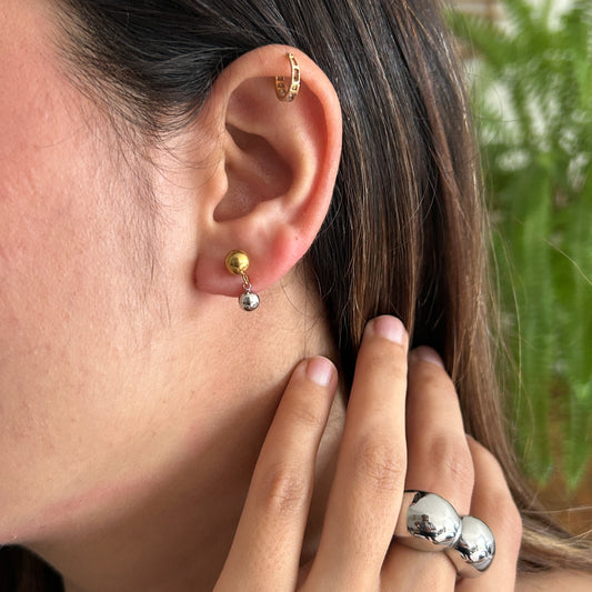 Close-up of a person's ear wearing a small gold hoop in the cartilage and a silver-and-gold dangle earring, with a large chunky silver ring on the hand.