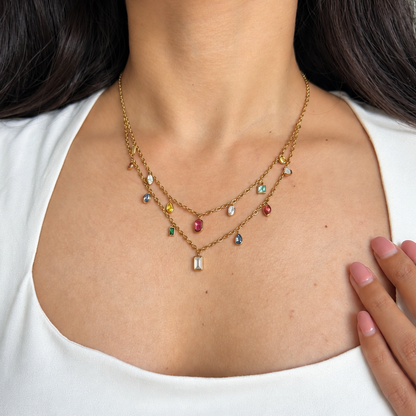 A close-up of a woman wearing a delicate gold double-layer necklace with small colorful gemstone accents and a central rectangular charm, paired with a white scoop-neck top.