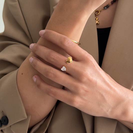 A close-up lifestyle shot of a model wearing a delicate gold ring with a small clear heart-shaped gemstone on her finger, paired with a beige blazer.