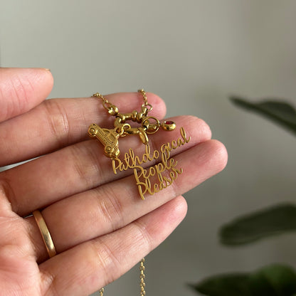 A gold necklace with a 'Pathological people pleaser' script pendant and a small animal charm, held in a person's hand against a soft-focus background.