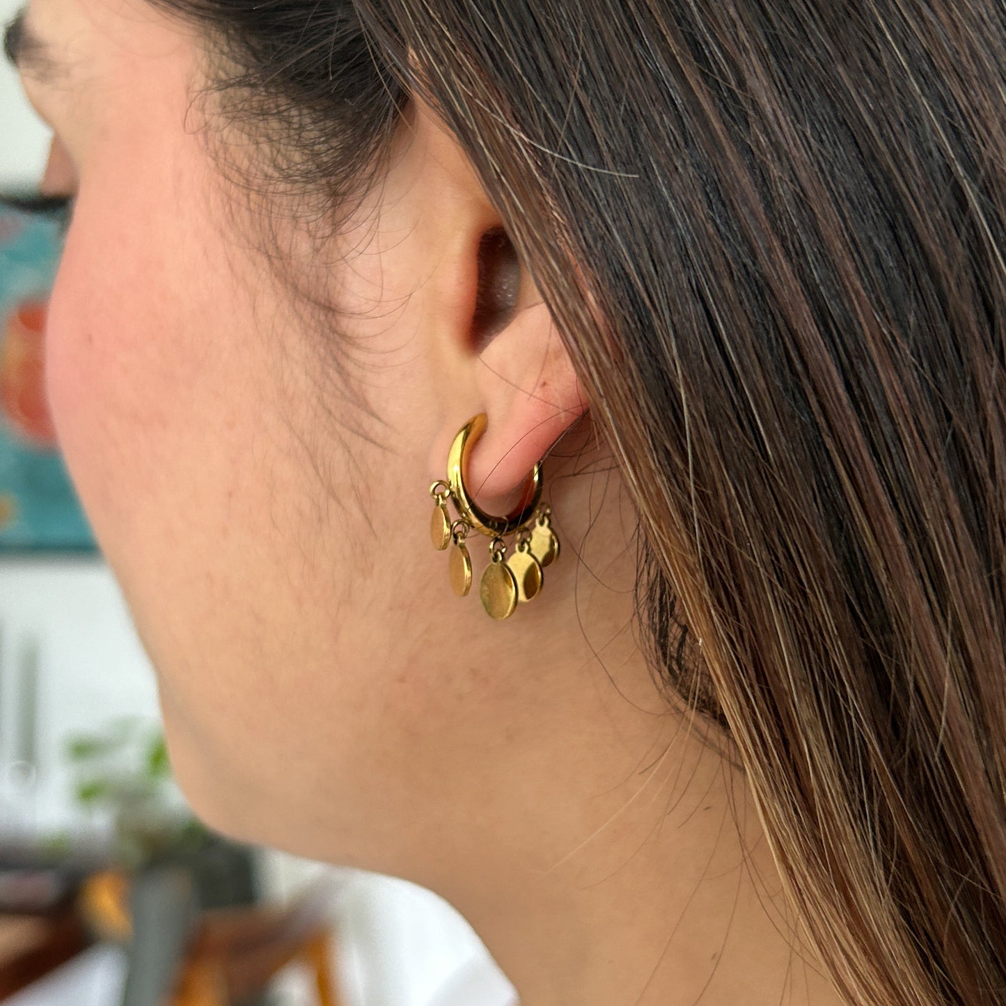 A close-up side profile of a woman wearing a small gold hoop earring adorned with dangling gold charms, set against a softly blurred indoor background.