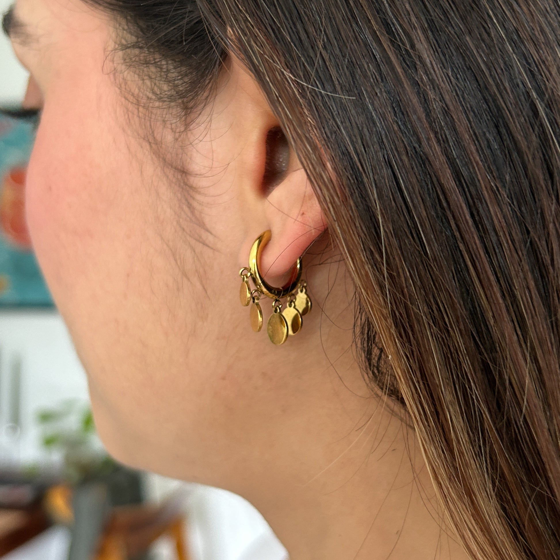 A close-up side profile of a woman wearing a small gold hoop earring adorned with dangling gold charms, set against a softly blurred indoor background.