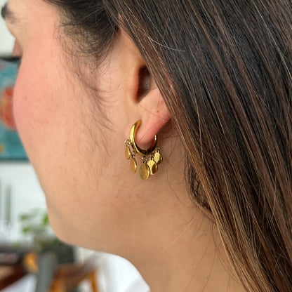 A close-up side profile of a woman wearing a small gold hoop earring adorned with dangling gold charms, set against a softly blurred indoor background.