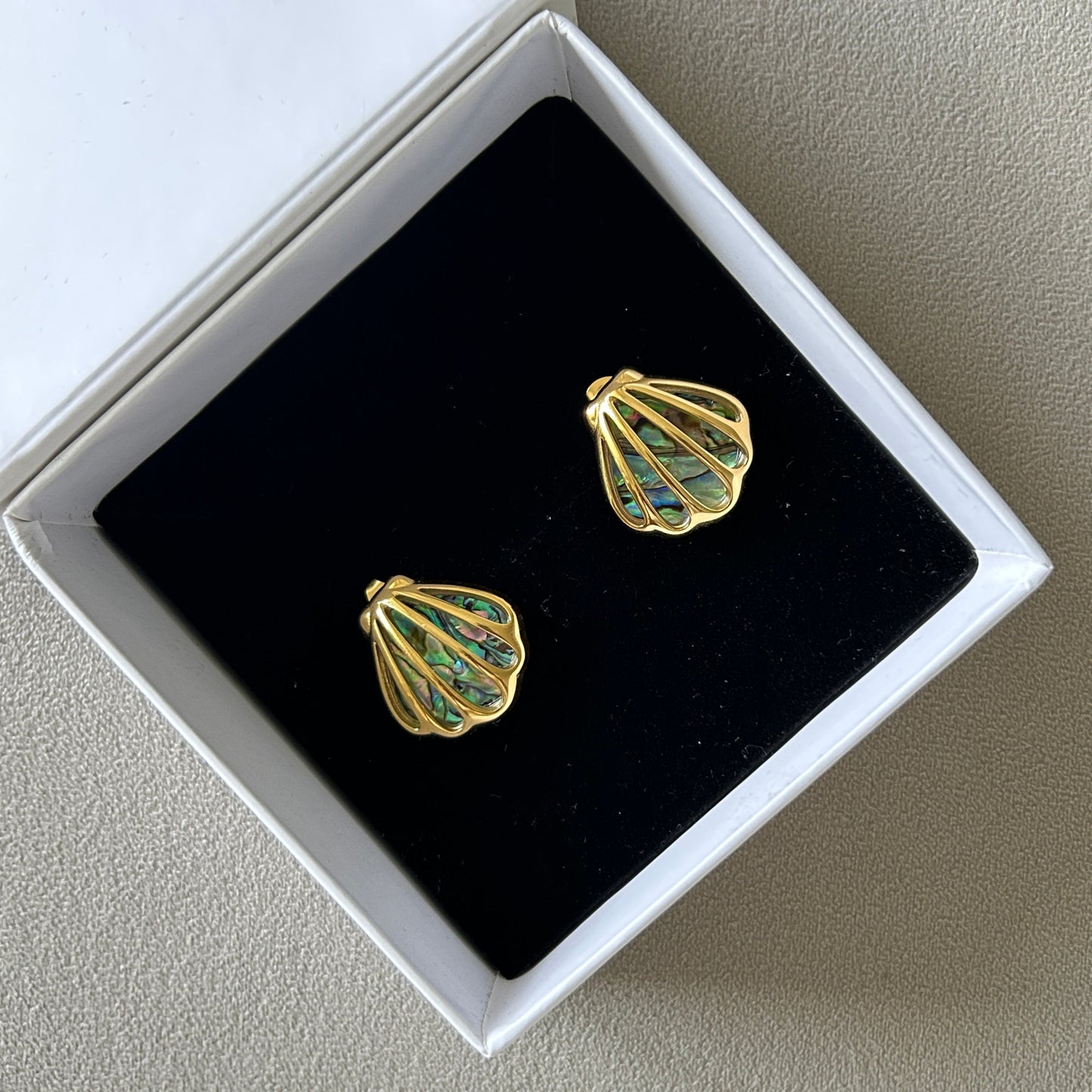 Gold and green shell-shaped mermaid earrings in a white box on a gray surface