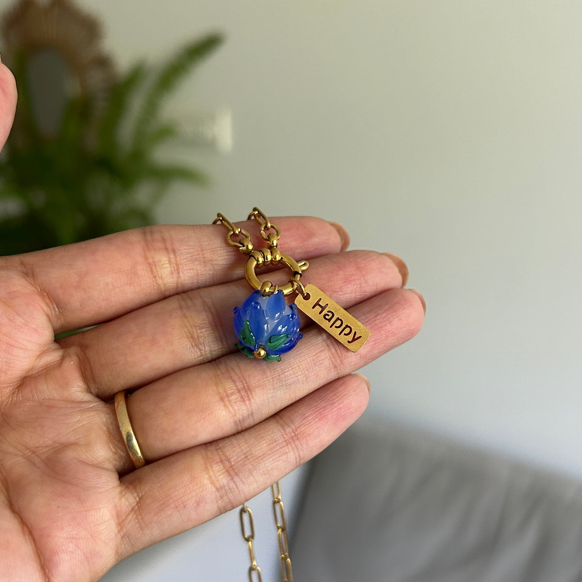 A gold chain necklace with a blue faceted bead charm and a small gold tag engraved with 'Happy', held in a person's hand against a blurred indoor background.