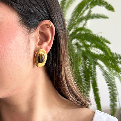 A close-up side profile of a woman wearing a chunky, gold-toned oval earring with a dark brown center, set against a soft-focus background of a white wall and green fern leaves.