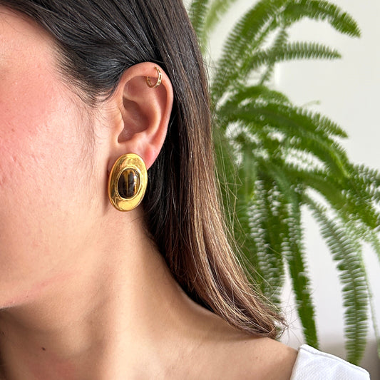 A close-up side profile of a woman wearing a chunky, gold-toned oval earring with a dark brown center, set against a soft-focus background of a white wall and green fern leaves.