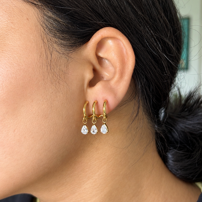 A close-up of a person's ear wearing a pair of gold hoop earrings with three small teardrop-shaped clear stones dangling from each hoop.