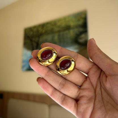A pair of gold-toned oval earrings with deep red cabochon stones, held in a person's hand against a softly blurred indoor background.
