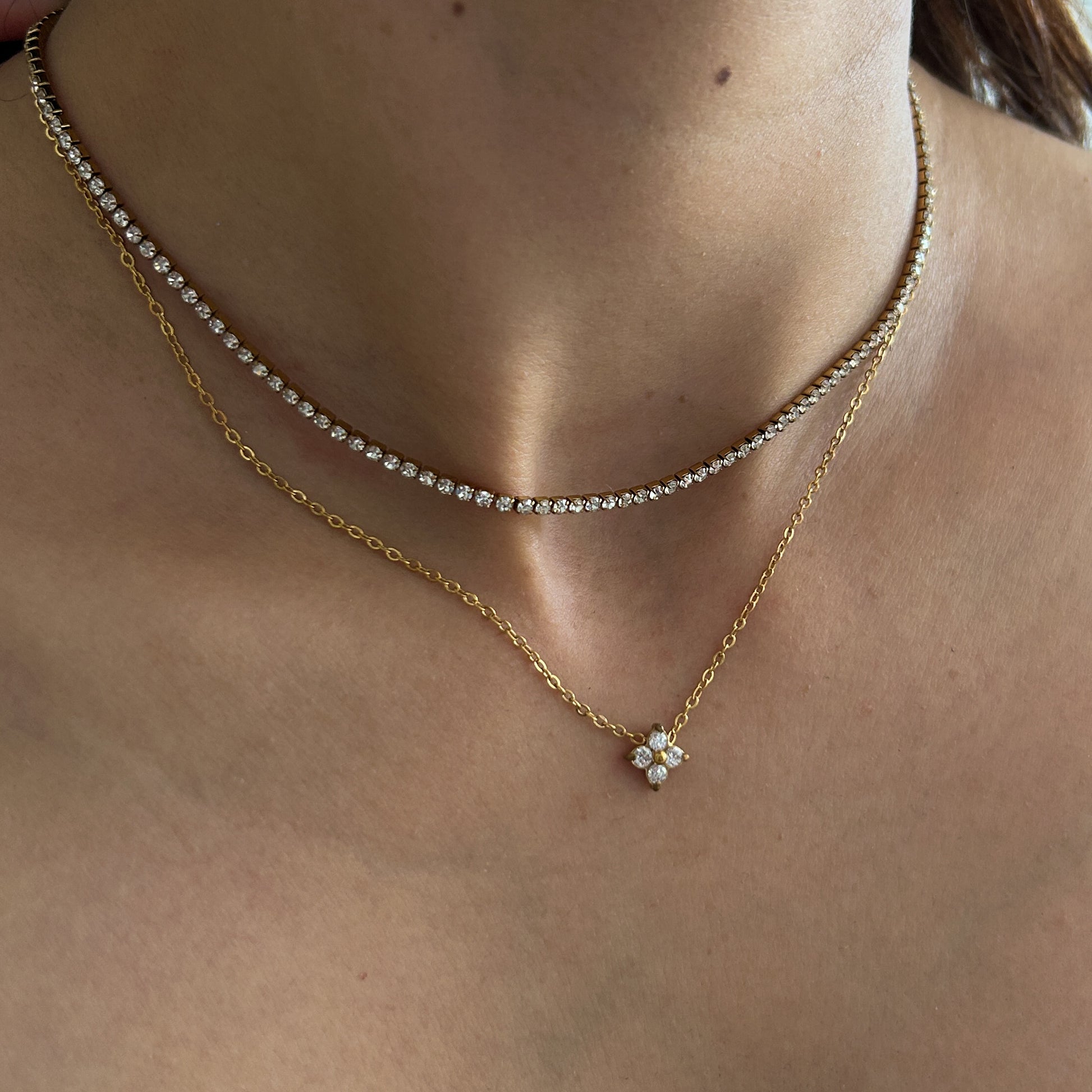 A close-up of a person's neck and upper chest wearing two layered gold necklaces: one delicate tennis-style chain with small clear stones and one longer chain with a small flower-shaped clear-stone pendant.