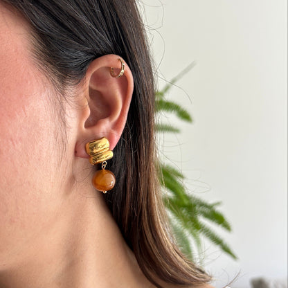 A close-up of a woman's ear wearing a gold-toned dangle earring with a textured top and a round amber-brown bead drop, against a softly blurred indoor background.