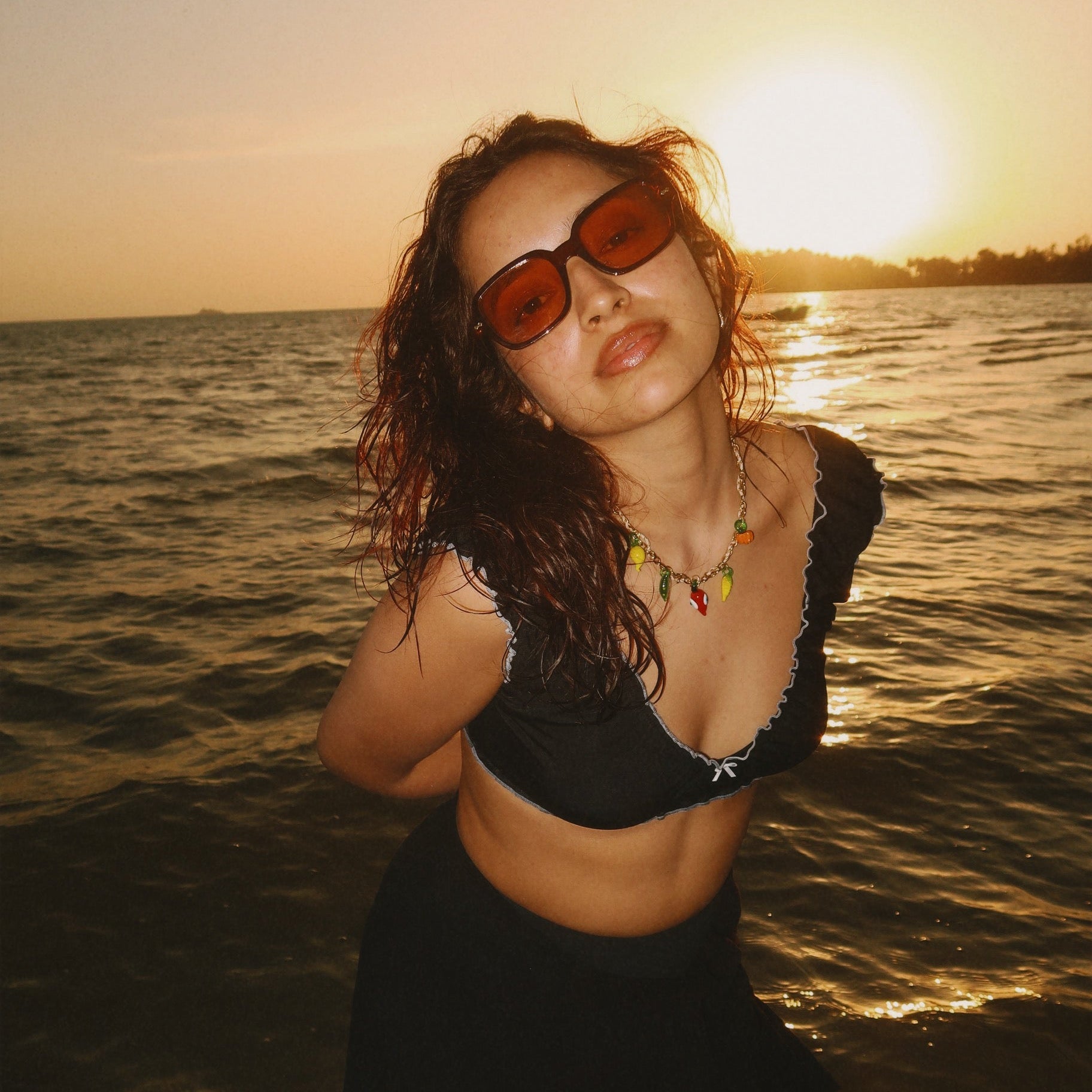 Woman in a black outfit with sunglasses standing in water at sunset wearing a fruit charm necklace by delta charms
