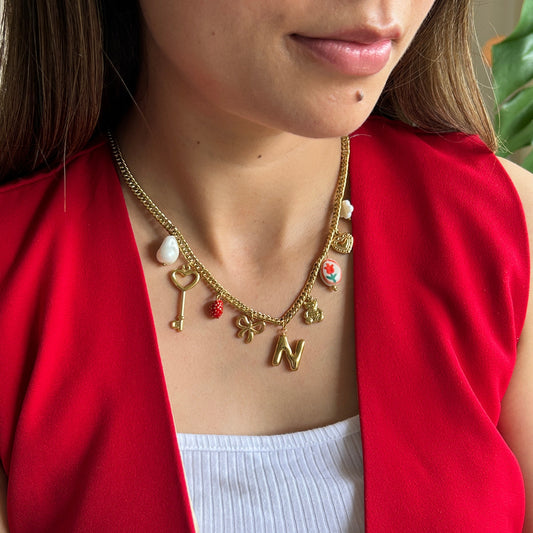 A close-up of a woman wearing a gold charm necklace with various letter and heart charms, styled with a red blazer over a white ribbed top.
