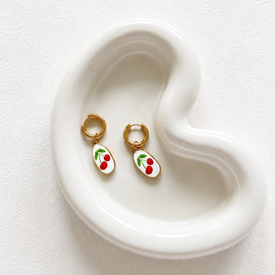 A pair of small gold hoop earrings with white oval charms decorated with red cherries and green leaves, displayed on a white ceramic heart-shaped tray against a textured white background.