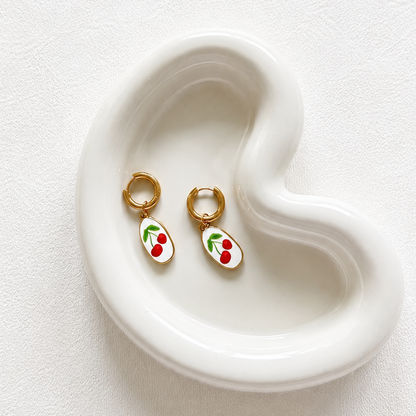 A pair of small gold hoop earrings with white oval charms decorated with red cherries and green leaves, displayed on a white ceramic heart-shaped tray against a textured white background.