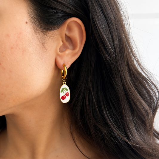 A close-up of a woman wearing a gold huggie hoop earring with a dangling white enamel charm featuring red cherries and green leaves.