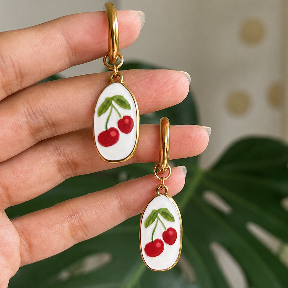 A hand holds a pair of gold-tone drop earrings with white oval charms decorated with red cherries and green leaves, set against a blurred background with a green plant leaf.
