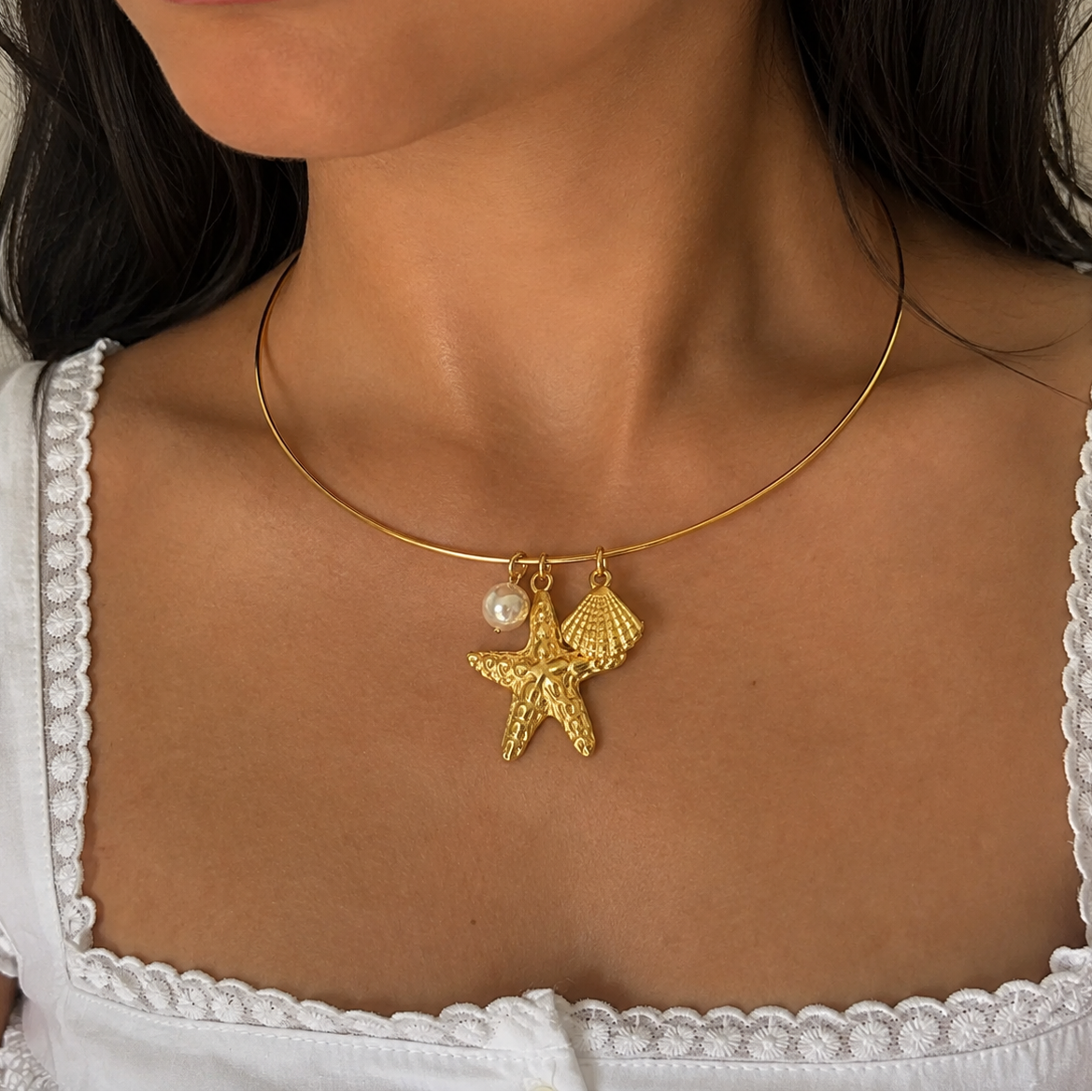A close-up of a model wearing a delicate gold necklace with a textured starfish and shell pendant, accented by a small white pearl, styled with a white lace-trimmed top.