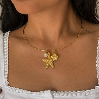 A close-up of a model wearing a delicate gold necklace with a textured starfish and shell pendant, accented by a small white pearl, styled with a white lace-trimmed top.