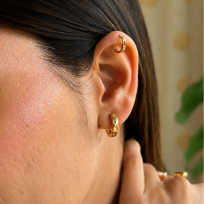 A close-up of a woman's ear wearing a small gold hoop earring and a gold cartilage piercing, with her hand near her neck.