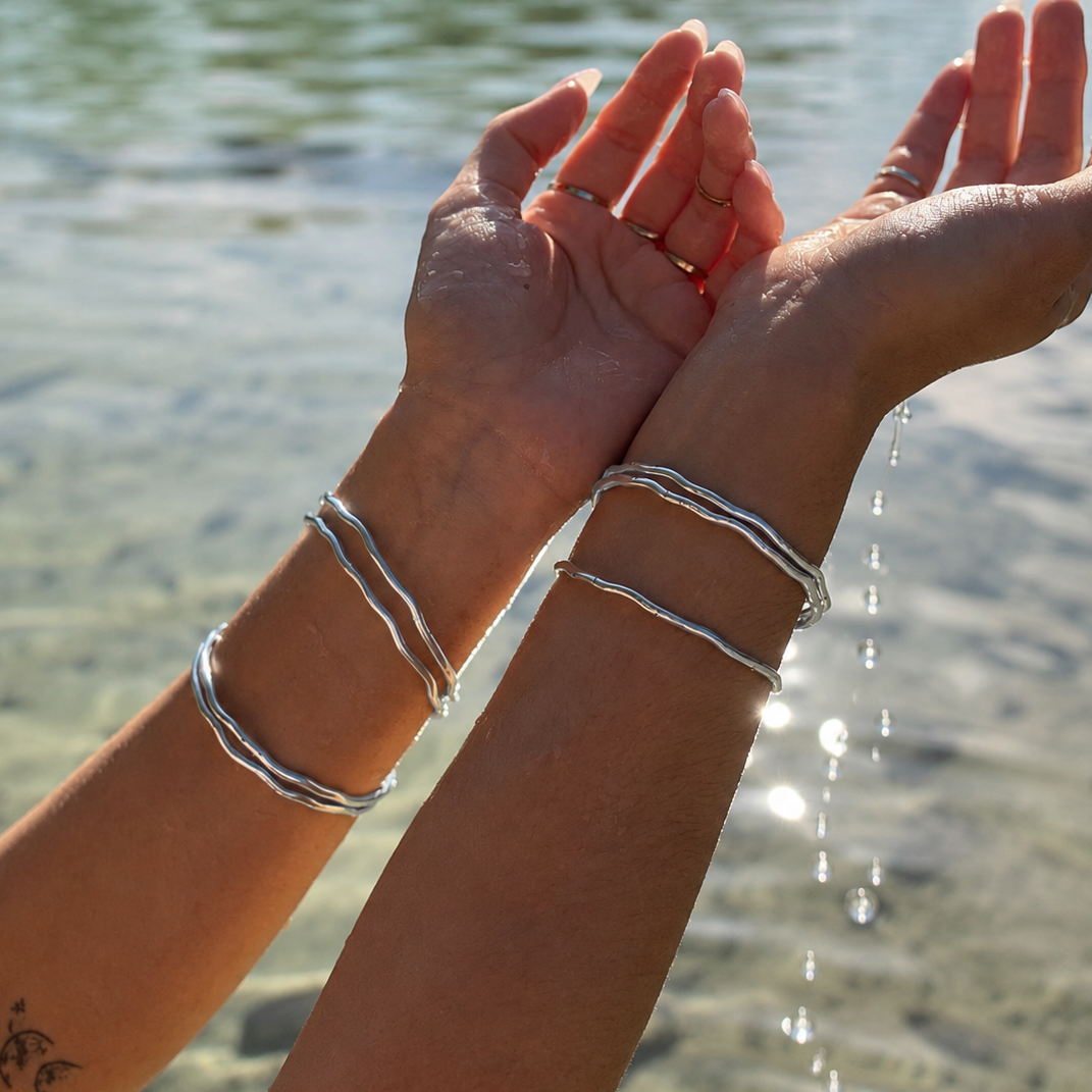 Two silver wavy double bangles worn on a person's wrists, shown with water dripping from their hands in an outdoor setting near a body of water.