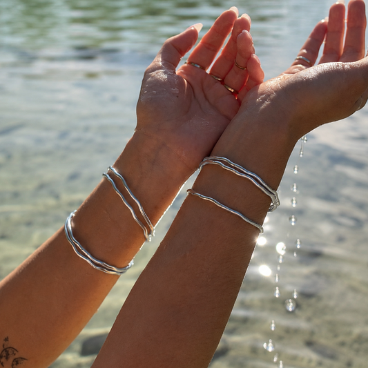 Two silver wavy double bangles worn on a person's wrists, shown with water dripping from their hands in an outdoor setting near a body of water.